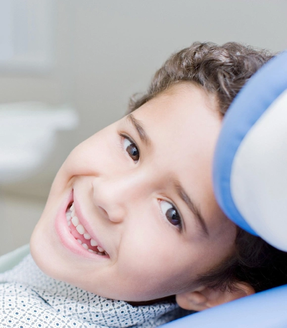 Young child smiling at dentist's office with blue dental equipment visible in background.