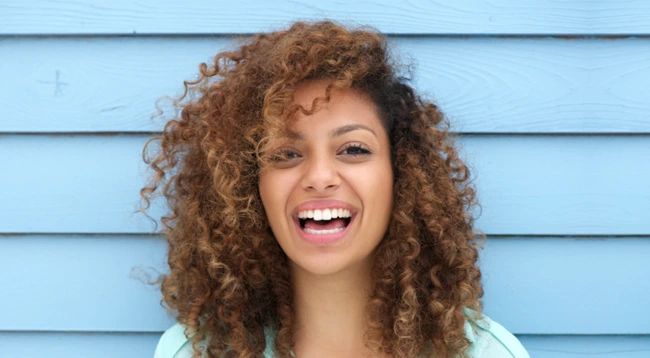 Smiling woman with curly auburn hair against blue horizontal siding background for dental practice.